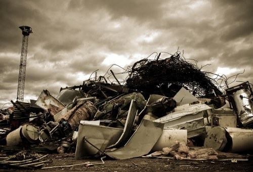 A large pile of mixed waste materials located outdoors on a flat surface, consisting primarily of white and grey household appliances and electronic waste, including washing machines, refrigerators, and metal parts, some of which are dented or damaged. The pile also contains tangled black wires, a large tangled mass of dark, twisted cables or debris, and smaller items like broken plastic components. The waste is stacked haphazardly, with some appliances partially concealed behind others, and some items leaning or resting on top of each other. In the background, there is a cloudy sky with a tower or pole to the left, and the scene appears to be in an open, industrial or disposal area near Wanstead in the UK, reflecting typical rubbish removal or waste collection activities conducted by companies like Commercial Waste Wanstead.