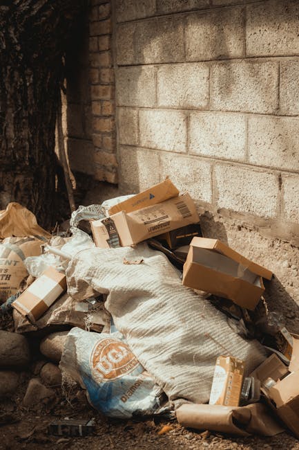A close-up view of an outdoor rubbish pile on a gravel surface, featuring a variety of waste materials including black plastic rubbish bags, discarded cardboard boxes, a yellow plastic crate, and a la