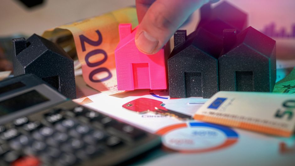 A person's finger is positioned near a row of small, black, house-shaped models made of textured material, arranged on a surface among loose Euro banknotes, including a visible 200 euro bill. In the f