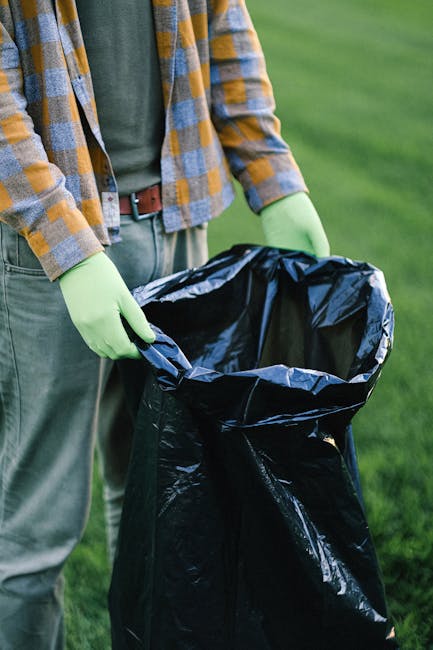 A person dressed in orange protective coveralls and wearing a white glove is holding a large blue plastic rubbish bag by its gathered top, which is tightly sealed. The bag appears to be filled with wa